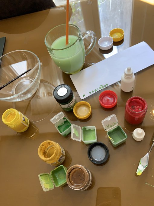 Various small jars and containers of paint in colors like yellow, green, red, white, and brown are scattered across a glass table, alongside a glass of green liquid with an orange straw, mixing palettes, and a paintbrush.