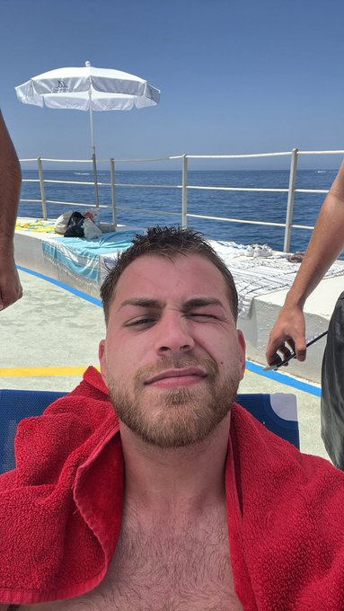 A man with a red towel over his shoulders squints into the sun on a boat deck, with the ocean and a white umbrella behind him—capturing a relaxed morning vibe that pairs well with a request to drop a $SOL address for tips.