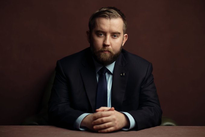The image features a man wearing a suit, sitting at a desk with his hands clasped together. The man appears to be looking directly at the camera, giving off an air of professionalism or perhaps contemplation. The background of the image is dark brown, which contrasts with the lighter colors in the foreground and adds depth to the scene. There are no other objects present in the image apart from the man sitting at the desk.
