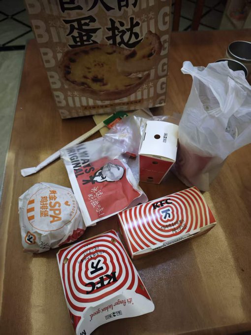 A tabletop holds assorted packaged snacks and a bagged drink, including red-striped KFC boxes, a round “鲜汁烧仙草” package, a “奶皮” item, and a large bag labeled “蛋挞.”