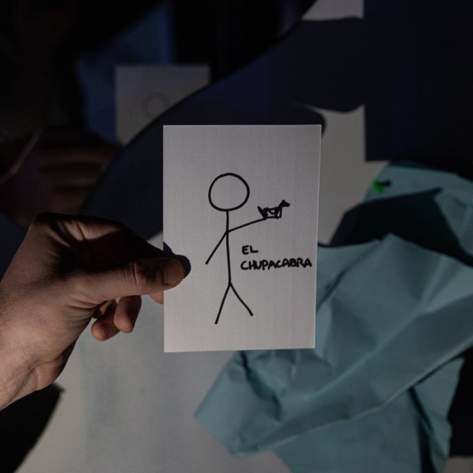 A hand holding up a simple stick figure drawing labeled 'EL CHUPACABRA' that depicts a person holding a small creature.
