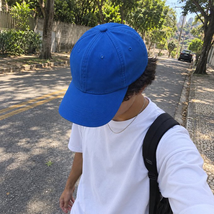 A young person wearing a bright blue baseball cap pulled low over their face, a plain white t-shirt, a thin silver chain necklace, and a black backpack strap, walks down a sunlit, tree-lined residential street with cobblestone curbs, likely hiding their identity while heading to or from their MEXC internship, embodying the relatable exhaustion of an 18-year-old who, despite having a professional title, is mostly just the target of their boss's endless demands to make things go viral.