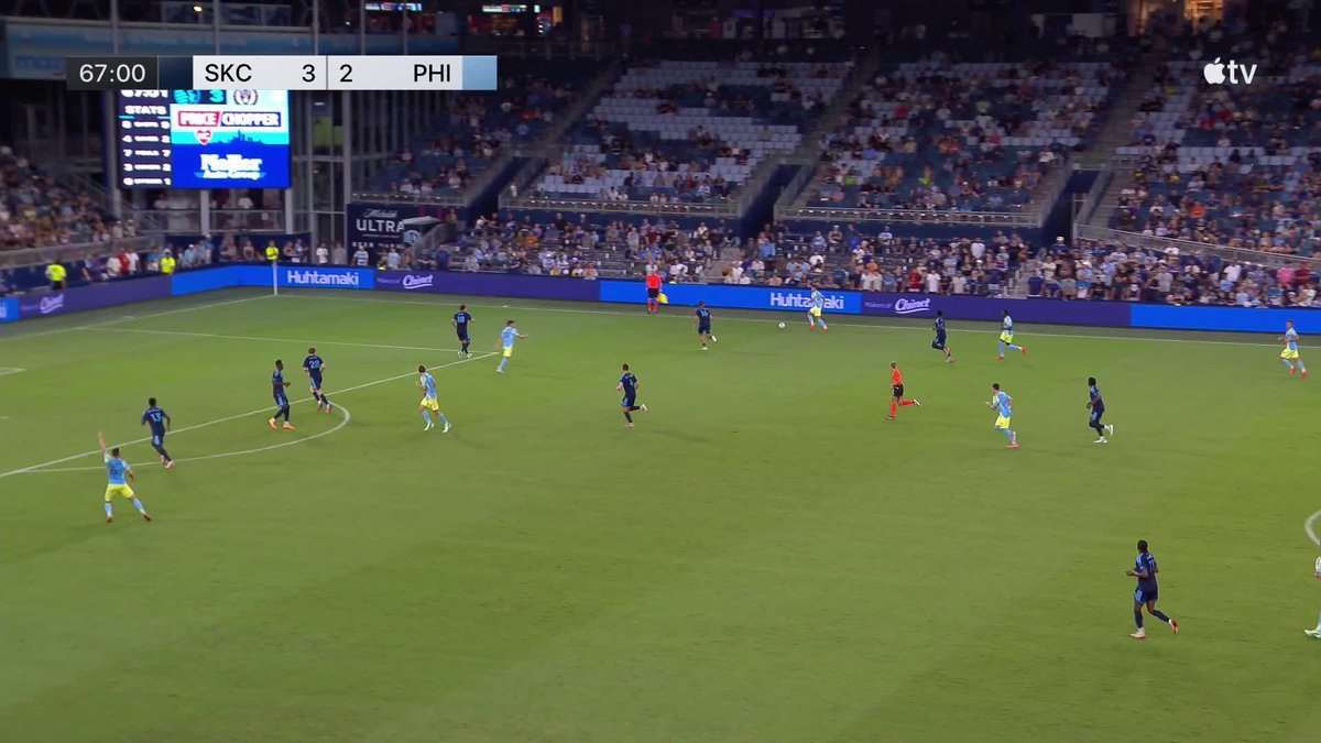 A soccer match is underway at night with the scoreboard showing "SKC 3 2 PHI" at 67:00.