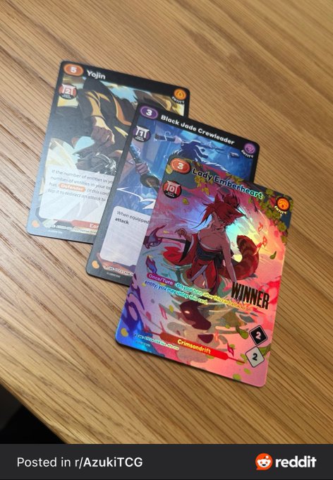 Three Azuki TCG cards are fanned out on a wooden table, featuring "Yojin" (cost 5), "Black Jade Crewleader" (cost 3), and the holographic "Lady Emberheart" (cost 3) foil alt art marked as the "WINNER" pull. The post celebrates the Azuki TCG launch day, and the image showcases exactly the kind of lucky pull the poster is wishing upon others—highlighting a sought-after holographic alt art card from the new set.