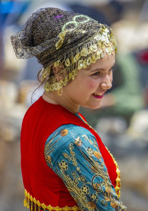 A young girl smiles while wearing an ornate traditional costume featuring a gold coin-decorated headdress, red velvet top, and intricately embroidered turquoise sleeves, suggesting cultural pride and artistry that aligns with the "QT Your Tezos Art" celebration of creative expression.