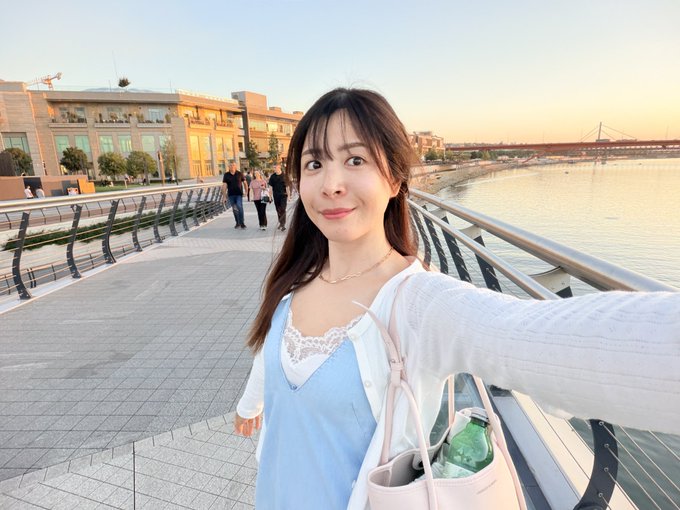 A woman in a light blue dress and white cardigan takes a selfie on a waterfront walkway with buildings and a bridge in the background.
