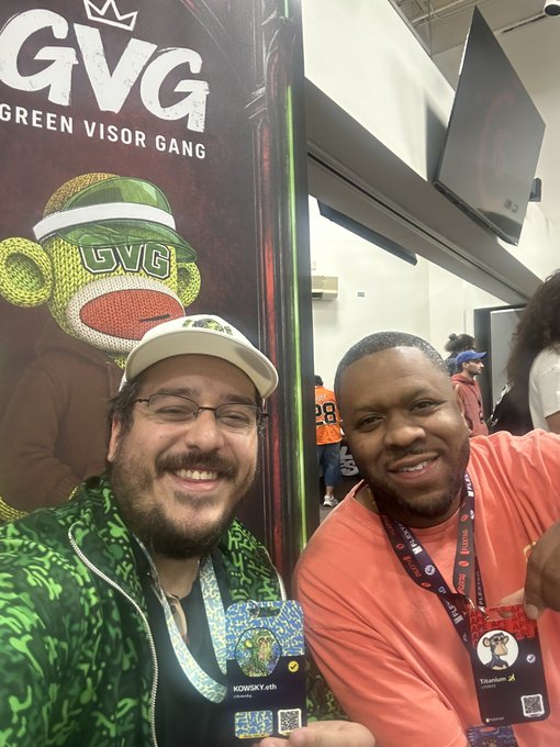 Two smiling men posing at an event, one in a green patterned jacket and glasses, the other in an orange shirt, standing in front of a banner that reads 'GVG GREEN VISOR GANG' featuring a cartoon monkey.