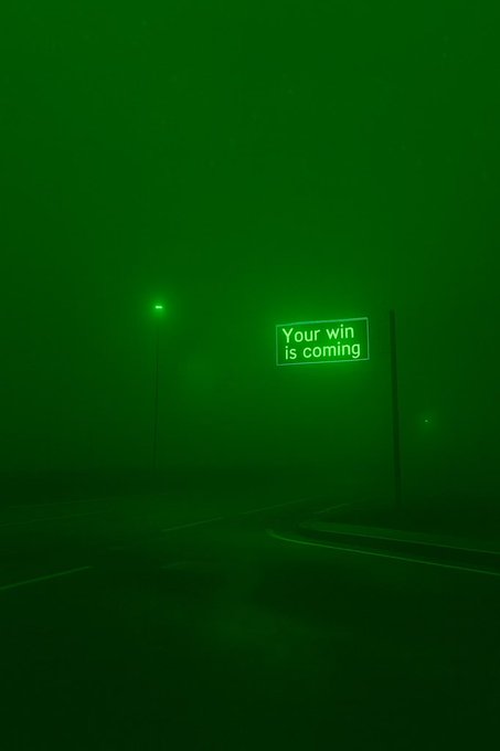 A dark, foggy road at night illuminated by a green streetlamp, with a glowing green sign reading "Your win is coming."