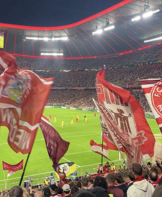 A view from the stands of Allianz Arena during a FC Bayern Munich match, with fans waving red flags as players in yellow jerseys compete on the pitch below, capturing the energetic stadium atmosphere. The image fits the "What's up fam?" post as a fan greeting from a thrilling night at the match.