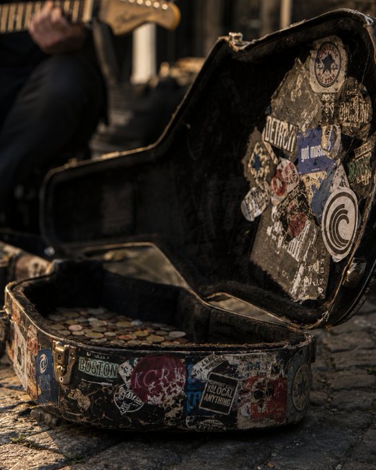 The image shows a guitar case with stickers, possibly from Detroit, placed on a cobblestone street or sidewalk. The case has been used as a makeshift storage container for various items such as coins and other small objects. The photo also includes text that reads "GM. Direct transfer. No intermediaries." This suggests that the person who owns this guitar case might have repurposed it to store their belongings, possibly due to limited resources or a desire to avoid using traditional storage solutions like banks or financial institutions.