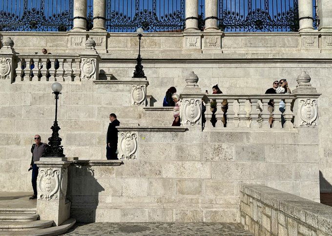 An outdoor architectural scene showing people gathered on and around the ornate white stone terraces and balustrades of a grand historic building under a clear blue sky.