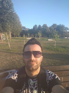 A man with dark hair, sunglasses, and a backpack takes a selfie outdoors in a sunny park setting with green fields and trees in the background, sharing a cheerful "Good Morning" vibe to match the post celebrating his art on @objktcom and @TransientLabs for #tezostuesday.