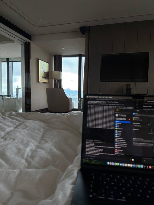 A laptop on an unmade hotel bed displays a trading interface with colorful market data and order books, set against a backdrop of a modern hotel room with floor-to-ceiling windows overlooking a misty cityscape—capturing the post's theme of deploying a trading bot from a remote location despite facing technical issues.