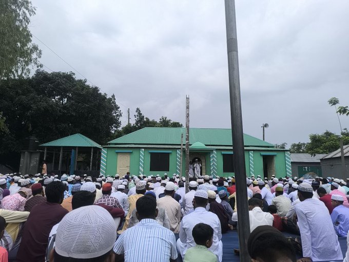 The image shows a large group of people gathered outside a green building, possibly attending an event or ceremony. The crowd consists of both men and women dressed in traditional clothing. A man stands at the front of the gathering, holding a flag that is part of a larger structure. The sky above them appears cloudy, suggesting an overcast day. The image also contains text elements such as "Good Morning All X fam GM Eid Mubarak to all my Muslim Brothers See you tomorrow with @NomismaNetwork and @useTria".