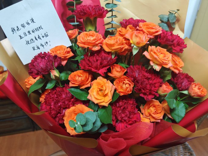 A vibrant bouquet of orange roses and red carnations wrapped in red and gold paper, accompanied by a handwritten Chinese note wishing the recipient a happy teacher's day.