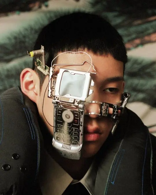 A person wearing a complex, futuristic metallic headpiece constructed from electronic components, a small screen, and wires covering half of their face, set against a textured bluish background.
