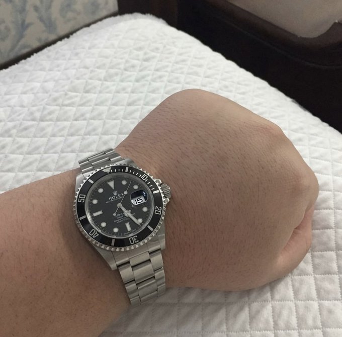 A wrist wearing a Rolex Submariner watch rests on a white quilted surface, showing the watch's black dial with the date "15" and "Rolex" branding clearly visible. The image directly relates to the post by showcasing the luxury timepiece being offered as a prize in the $0.01 giveaway contest.