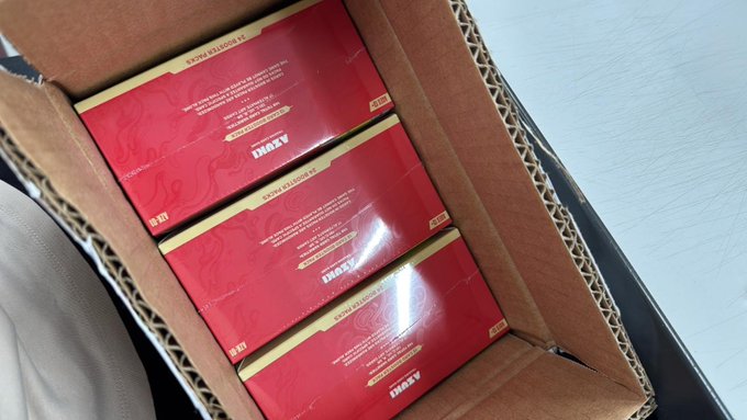 A cardboard shipping box holds several sealed red-and-gold "Azuki" trading card game booster boxes. The collector has just received their shipment and is showing off the unopened packs before revealing the contents.