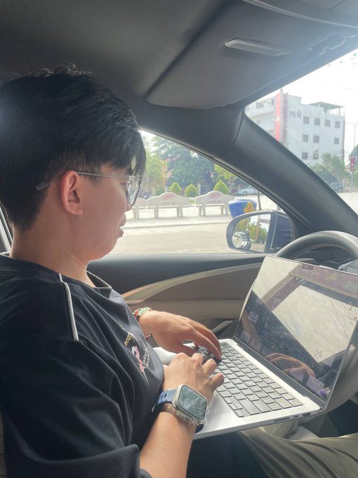 A young man wearing glasses and a smartwatch sits in a car, typing on a silver laptop resting on his lap.