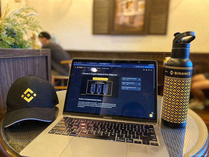 A laptop displaying a Binance Academy course titled "Course 2: Crypto Safety & Due Diligence" sits on a table alongside a Binance-branded water bottle and cap. The setup aligns with the post's mention of learning with Binance Academy, showcasing an educational session on crypto safety and due diligence.