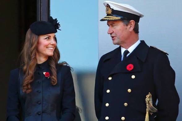 A smiling woman in a black fascinator and dark coat with a red poppy pin stands beside a man in a naval officer's uniform, also wearing a red poppy, as they exchange a warm glance. The image reflects the "daily smile" caption by capturing a tender, joyful moment between the two well-dressed individuals.