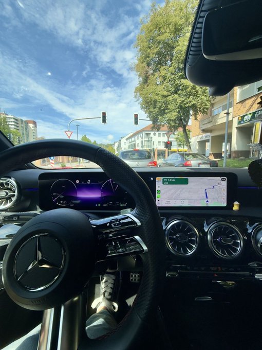 The image shows the interior of a Mercedes-Benz with the steering wheel logo clearly visible, a digital dashboard displaying navigation with "1.5 km" to the next turn, and a view through the windshield of a sunny street with traffic lights and trees. The caption relates to the post's casual "Good morning, Happy Wednesday" greeting by capturing a bright, pleasant morning commute scene from behind the wheel.