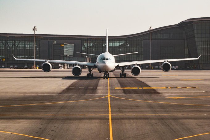 A large white commercial airplane with four engines sits on a tarmac in front of a modern, curved airport terminal building.