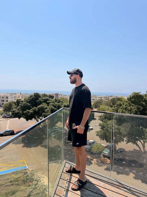A man in a black t-shirt, shorts, and cap stands on a glass-railed balcony overlooking a sunny coastal town, gazing out toward the sea — a relaxed "Gm friends" vibe matching the bright morning views below.