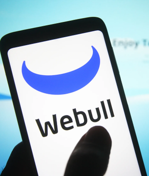 A smartphone displays the Webull logo against a blue background. The image illustrates the report that Webull will offer Bitcoin perpetual contracts to its more than 6 million clients.