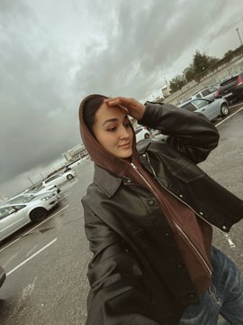 A young woman wearing a brown headscarf and black leather jacket shields her eyes from the sun in a parking lot on a cloudy day.
