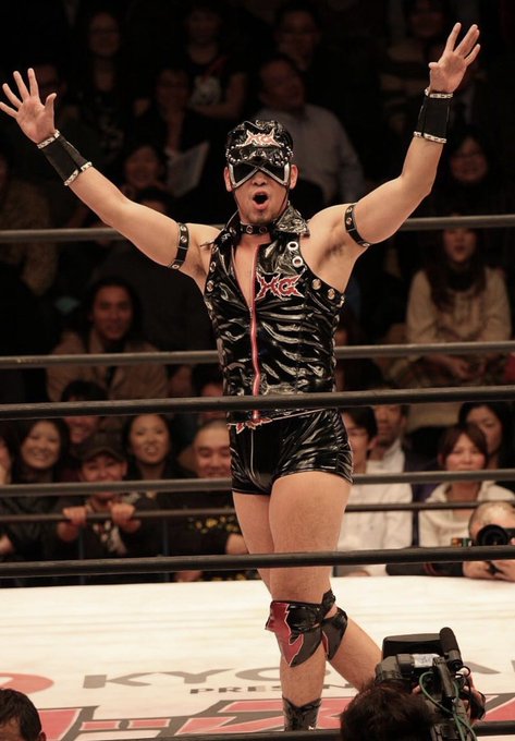 A professional wrestler wearing a black mask, black and red gear, and wrist guards stands in a wrestling ring with arms raised triumphantly while an audience watches from the background.
