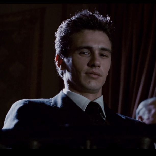 The image features a man wearing a suit and tie sitting at a desk with his eyes closed. The background of the image shows a dark room or office setting, which could be an office cubicle or a dimly lit room. There are two other people in the scene, one on the left side and another person partially visible on the right side. The man's gaze is directed off to the side, giving him a serious expression as he looks into the distance.
