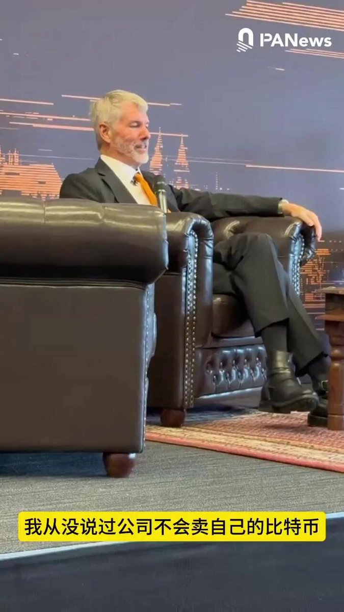 The image features a man sitting in a chair, wearing a suit and tie. The background of the photo shows a city skyline with buildings at night. There are two chairs visible in the scene - one where the man is seated and another that appears empty. A caption below the image reads "渣男就是这样： 我是说你不要卖 万 $BTC 没说过我不能卖 买种肉结".