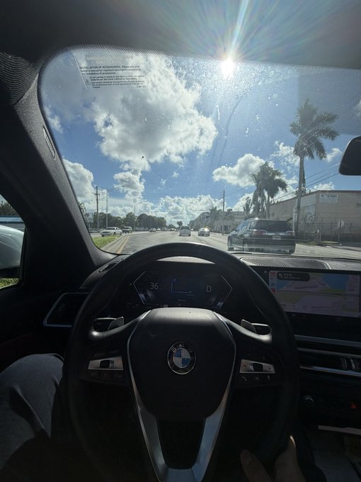 A driver enjoys the sunny afternoon from behind the wheel of a BMW, cruising down a palm-lined street with the digital dashboard showing "36" and "D" on the speedometer. The bright, carefree drive perfectly captures the post's message of savoring the weekend while staying focused.