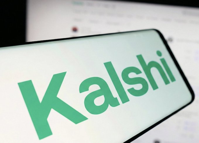 The image features a cell phone with the word "KALSHI" written on its screen, which is green. The background of the photo shows a computer monitor displaying a website or social media page. The text on the cell phone reads "BULLISH: KALSHI just picked SUI for its next wave of U.S. crypto derivatives". This suggests that the person who owns the cell phone might be interested in cryptocurrency and is using it to research, learn about, or invest in digital assets like cryptocurrencies. The website displayed on the computer monitor could provide information related to this topic, such as news articles, forums, or educational resources.