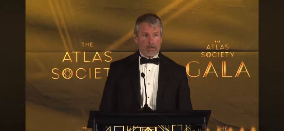 The image features an older man wearing a tuxedo standing at a podium with a microphone, giving a speech or presentation. He appears to be addressing an audience, possibly at a formal event like a gala. The setting includes a stage backdrop that is predominantly yellow and black in color, which contrasts with the gray wall behind him. The text "The Atlas Society Gala" can also be seen on the image.