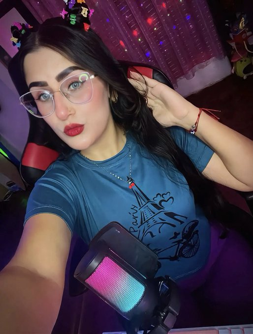 A young woman with glasses and dark hair, wearing a blue graphic t-shirt, sitting in a gaming chair with colorful RGB lighting in the background.