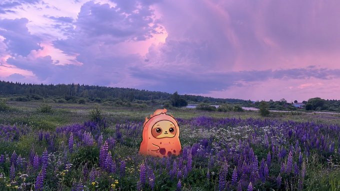 An adorable orange cartoon character with a smiling face stands in a purple lupine field beneath a dramatic pink and purple sunset sky.