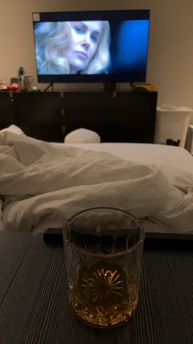 A dimly lit hotel or bedroom scene featuring a glass of amber-colored liquor on a dark table in the foreground, with rumpled white bed sheets, a dresser, and a TV playing a blonde woman's image in the background.