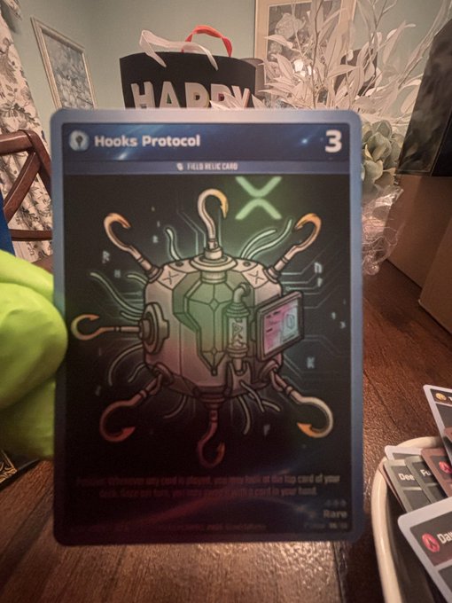 A close-up photo shows someone holding a holographic "Hooks Protocol" trading card (value 3) featuring a circuit-like robotic design with hook appendages, with a "Happy Holidays" gift bag visible in the background. The card relates to the post as the collector excitedly displays this rare Evernode/Xahau blockchain-themed card.