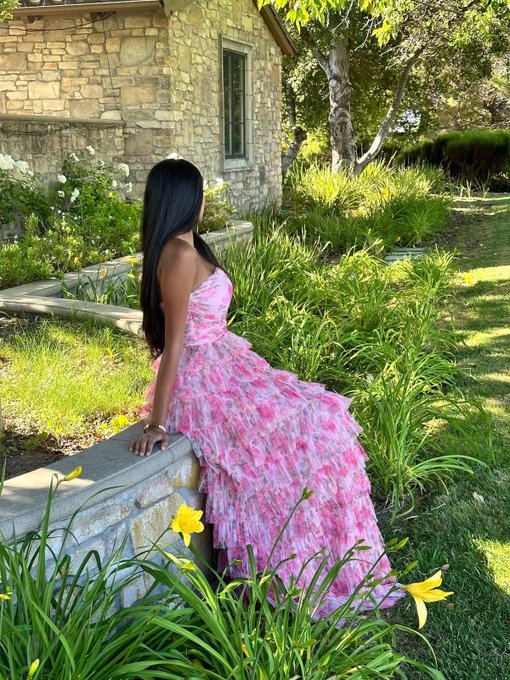 A woman with long dark hair wearing a flowing pink floral dress sits on a stone ledge in a lush green garden with yellow flowers near a stone building.