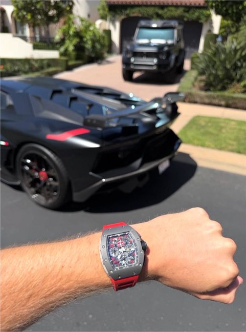 A wrist sporting a Richard Mille watch with a red strap is held up in the foreground, with a sleek black Lamborghini and a Land Rover Defender parked in the driveway of a suburban home in the background. The image visually reinforces the post's boastful tone of crypto success, using luxury cars and a high-end timepiece to symbolize wealth and winning status.