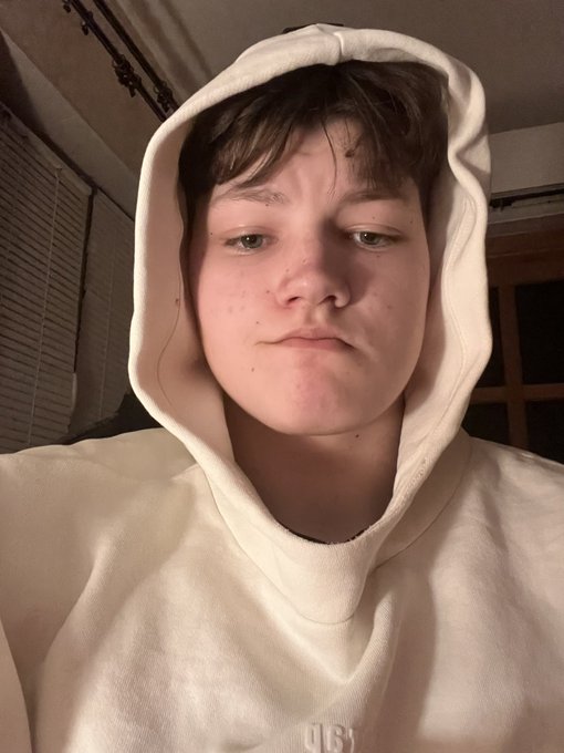 A person wearing a cream-colored hoodie takes a close-up selfie with a neutral expression in dim lighting.
