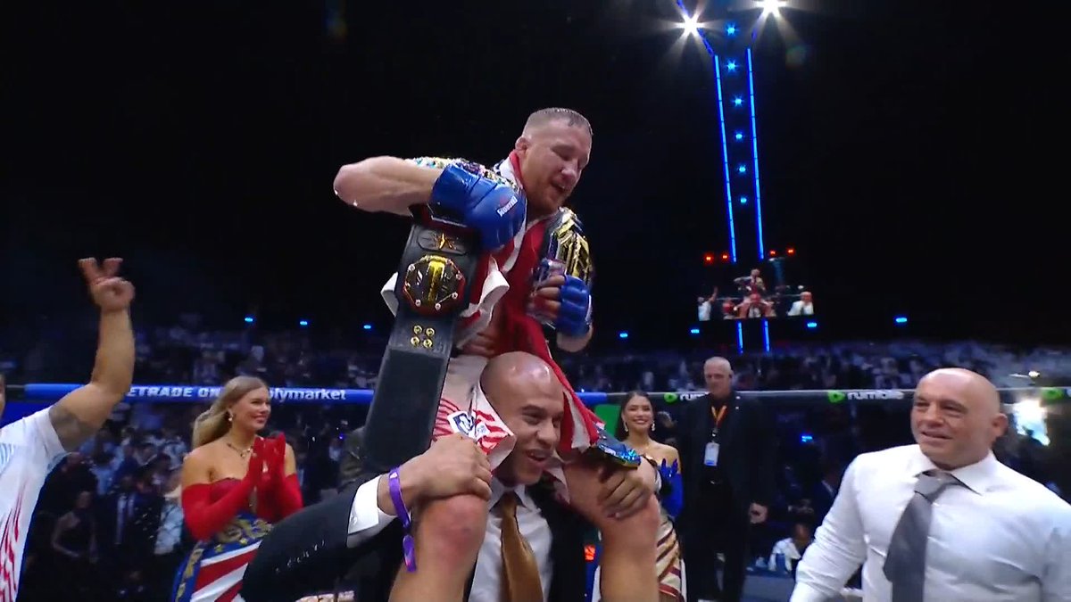 The image shows a man being lifted into the air, with his arms raised high above his head. He is wearing boxing gloves and appears to be celebrating his victory over another fighter. The caption of the image reads "The Highlight @Justin_Gaethje's the undisputed lightweight champion of the world! #UFCWhiteHouse [ Title fights presented by @Cryptocom ]". This suggests that the man being lifted into the air is a heavyweight boxing champion, and he has just won his title in the fight.