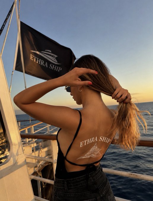 A woman on a boat at sunset gathering her hair, revealing an 'ETHRA SHIP' tattoo on her back matching the boat's black flag logo.