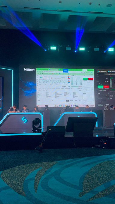 A view of a crypto trading event stage featuring the 'Bitget' logo, with a large screen displaying trading charts and a panel of people seated at a desk.