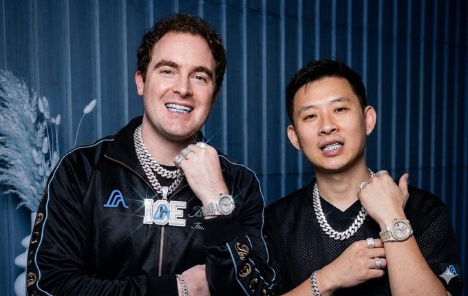 Two men posing together wearing matching black 'ICE' jackets and thick diamond chains; the man on the left has curly hair and the man on the right has dark hair, both sporting diamond jewelry.