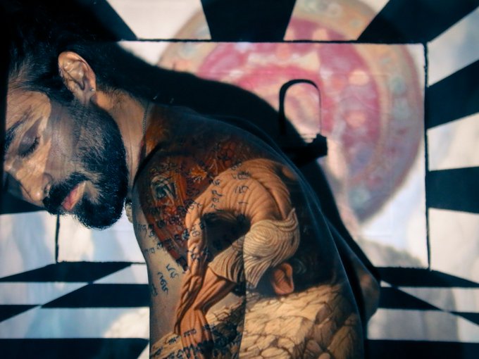 Dramatic photograph of a bearded man with extensive muscular and script tattoos on his back, set against a black and white checkered background.