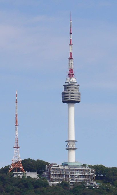A tall red and white telecommunications tower with an observation deck stands prominently against a blue sky, with a smaller transmission tower visible to the left.