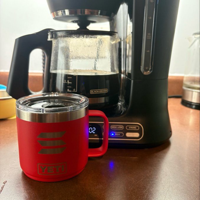 A red YETI insulated mug sits on a wooden surface in front of a Black+Decker coffee maker actively brewing coffee.